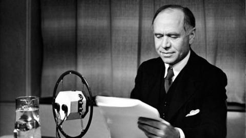 Lord Beaverbrook looks at script, behind microphone and glass of water.