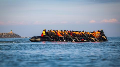 Migrants wearing orange lifevests sit on an inflatable boat on the English Channel, a rocky headland appears in the backdrop