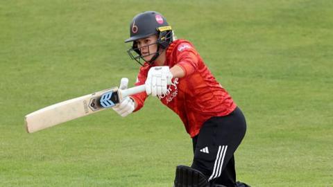 Lucy Higham of The Blaze plays a shot as Nat Wraith of Warwickshire Bears looks on during the Vitality Blast Women match between The Blaze and Warwickshire Bears