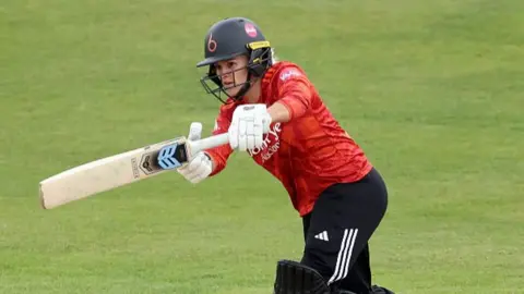 Lucy Higham of The Blaze plays a shot as Nat Wraith of Warwickshire Bears looks on during the Vitality Blast Women match between The Blaze and Warwickshire Bears