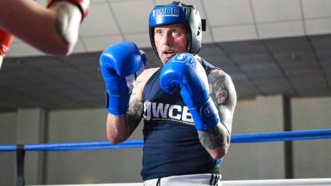 A man wearing boxing attire, including a blue helmet and gloves, stands in a boxing ring with his hands raised ready for a fight. He is wearing a dark blue sleeveless shirt and there are tattoos covering both his arms.