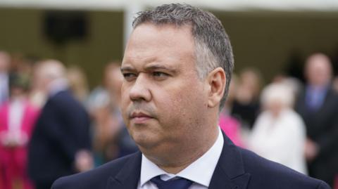 A man dressed in a navy suit and tie with a white shirt looks to the left of the camera. He is clean shaven and has short, dark hair which is turning grey at the sides. Behind him is a crowd of people in formal clothing, which is out of focus. 