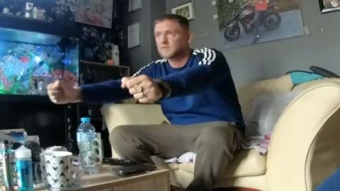 A man sat on a sofa with his arms in front of him. There is a photo behind him on the wall, a fish tank to his right and bottles and glasses on the table.