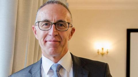 Lloyd Cooke wears glasses, a dark grey suit jacket, white shirt and pale blue paterned tie. He is smiling at the camera.