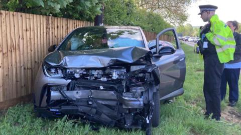 A damaged car with a crushed front end has come to rest beside a wooden fence and grassy verge, while a police officer and a woman stand nearby assessing the scene along a roadside lined with trees.