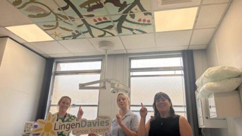 Three women are standing in a hospital room and are pointing at the ceiling. On the ceiling there is a hand-painted mural which has green plants, brown trees, colourful leaves, bugs and a bird painted on it