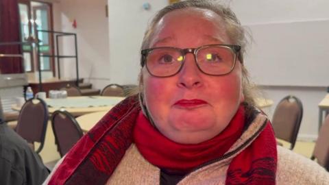 Jay Lander, who is wearing a cream jumper and red scarf, is sitting on a chair during a choir singing session. 