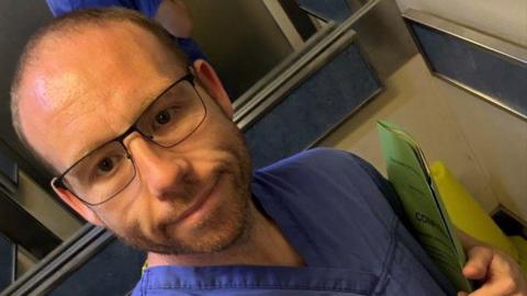 A smiling slim faced male midwife (Lee Gigg) wearing; glasses, blue hospital scrubs, carrying a green medical records file .