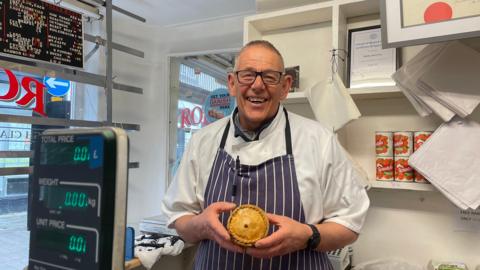 Paul Jessop, wearing butcher's apron, standing in his shop, holding a pork pie