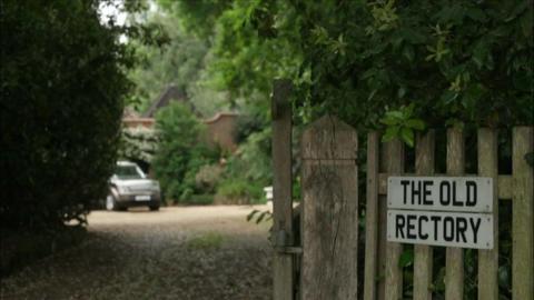 A sign on a wooden fence that is made out of white car number plates and reads "The Old Rectory". There is a driveway to the left of the fence and a car is parked at the bottom of the drive. 