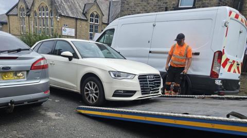 A cream-coloured Audi car is loaded onto the back of a lorry, while an official in orange hi-vis clothing supervises. There is a grey car to the left, parked on the road, with a white van behind the action, parked on the other side.