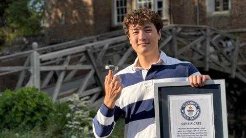 A young man is standing outdoors in front of a wooden, arched bridge that crosses a river, with an old college-style building visible behind it. He has short, curly brown hair and is wearing a blue-and-white striped rugby shirt. With his right hand, he is balancing a fidget spinner on one finger, demonstrating control and focus. In his left hand, he holds a Guinness World Record certificate. The setting has a calm, historic university atmosphere, likely on a campus.
