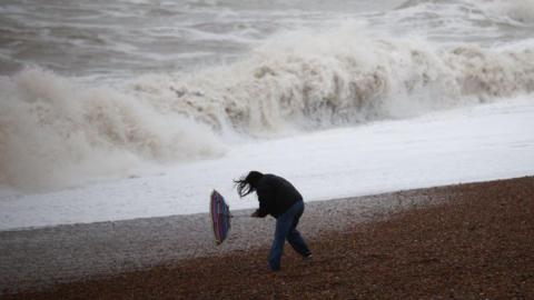 A woman holds on to her broken umbrella as she has walks along a beach in stormy weather 