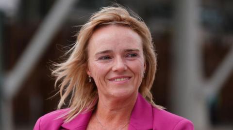 Nurse Sandie Peggie has short blonde hair and wears a pink blazer and smiles looking off to the side.