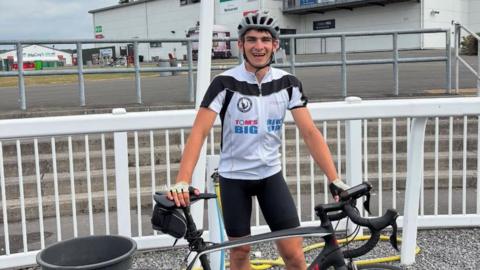A smiling Tom Dunlop standing outside a racecourse wearing cycling gear and holding his bike.