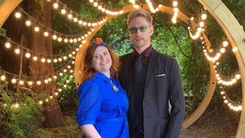 Kelly Stuart and Nick Wilson. Kelly has red hair and wears a blue dress, while Nick is blonde and wears a suit. They are surrounded by trees and a string of lightbulbs is draped around a wooden structure behind them.