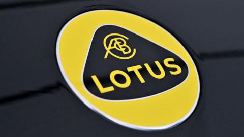 The Lotus logo. It has yellow writing on a black background surrounded by a triangular, curved shape. That is then surrounded by a yellow circle. The logo sits on a black, metallic surface.