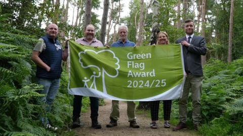 A group of four men and a woman stand in a wood surrounded by trees. They are holding up a green and white flag, with a white tree logo and text reading "Green Flag Award 2024-25"