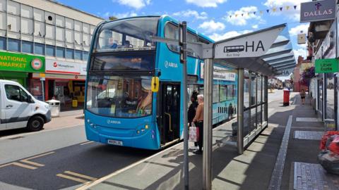 A blue Arriva bus waiting at a bus stop in Leicester with two passengers queuing to get on 