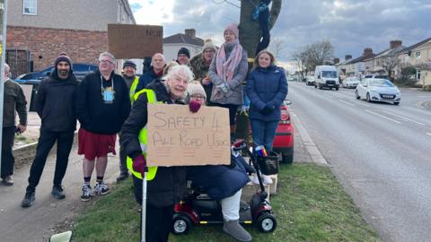 A group of campaigners stand in front of a tree on a residential road. Two of them hold protest signs on brown cardboard - the front one says 'safety 4 all road users'. Knitted items are attached to the trunk of the tree.