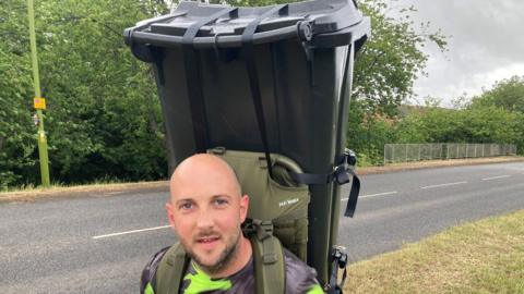 Daniel Fairbrother, with a large wheelie bin on his back. He is looking at the camera, wearing a colourful T-shirt. He is by a road with a grass verge, and there are trees on the other side. 