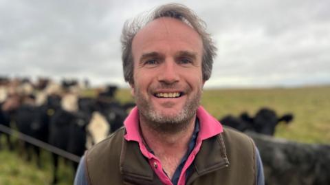Johnnie Balfour wearing a brown gilet with a a blue jumper and a pink underneath. He is standing in front of a field of cows
