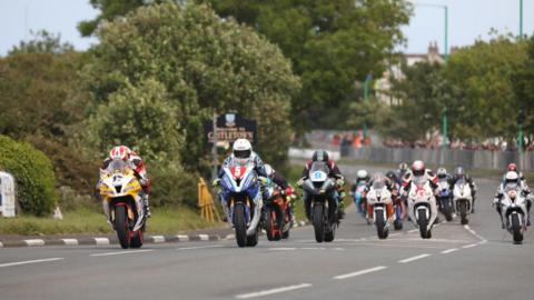 A large group of motorbikes racing on the Billown Course. The bikes and riders' leathers are various colours and there are spectators watchingfrom behind a barrier in the background.