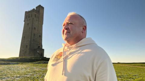 A man wearing a cream-coloured hooded top is standing in front of Castle Hill in Huddersfield, which is a large castle-like tower on a hillside. There is frost on the ground and a clear blue sky. He is looking out into the distance.