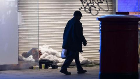 A stock image of a homeless person sleeping in a shop doorway with a silhoutted man walking past him.