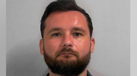 A police custody image of a man with short dark-brown hair, brown eyes and a trimmed brown beard. He has a serious expression as he looks into the camera against a plain light-grey backdrop. He is wearing a blue and white plaid shirt over a white t-shirt.