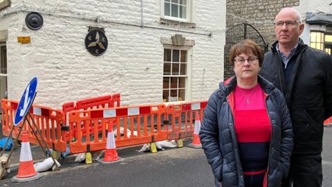 A man in a black coat with short grey hair and wearing glasses and a shorter woman with brown hair, wearing glasses, a blue coat and pink and red top stand in front of red plastic barriers protecting a hole in the pavement outside a white-painted stone building.