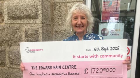 A woman stands in front of a granite building - she holds a giant cheque for £172090.00 from the National Lottery. She is smiling 
