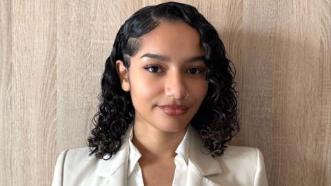 Mel Therese poses for the camera. She has curly, shoulder-length black hair and is wearing a white suit.