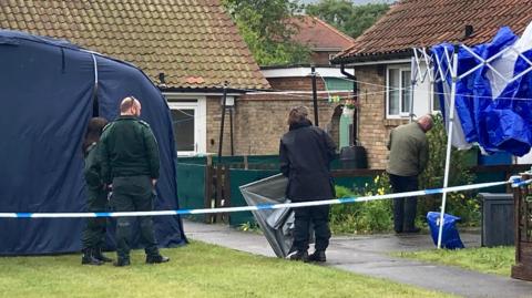 Emergency service workers stand on the grass beside two yellow brick bungalows. There are two officers to the left in dark green trousers and jumpers, and a third to their right in a black coat and trousers, all with their backs to the camera. A fourth person stands on the path to a house in a khaki green jacket. There is a navy tent to the left and a blue and white gazebo to the right, with blue police tape running across the front of the area.