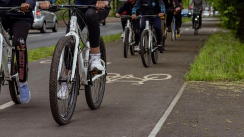 A group of children dressed in dark clothing cycling on white mountain bike on a cycle path beside a busy road