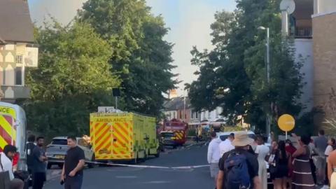 An emergency services scene on a residential London street cordoned off with police tape. A large yellow ambulance with red chevron markings is parked in the road, with fire engines visible in the background and a crowd of onlookers gathered on the pavement.
