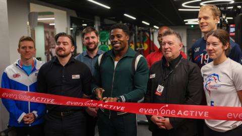 A group of eight athletes, including Harry Aikines-Aryeety, standing behind a red ribbon branded with white "Everyone Active" branding. They are smiling for the camera as Aikines-Aryeety holds scissors open, ready to cut the ribbon.