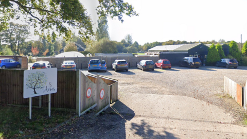 The entrance of 8 Acres care home. A sign with a tree logo is by a fence and cars are parked by multiple buildings.