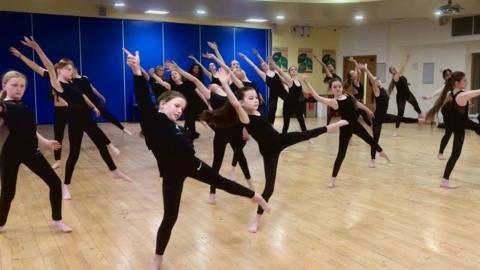 Children in a dance lesson and the are in the middle of doing a dance move. They are all wearing all black.