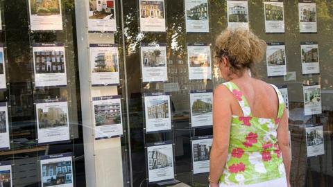 Woman in floral sleeveless top looks in estate agent window at rows of listings