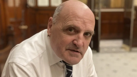 Steve Radford, who is bald and is wearing a white shirt and tie, sits in Liverpool Town Hall 