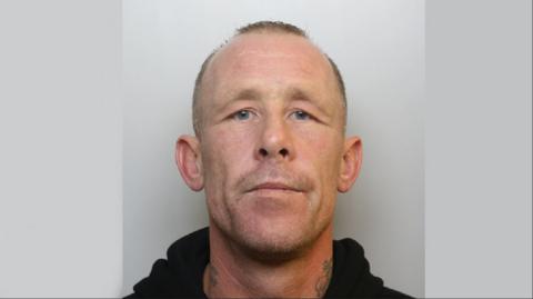 A mugshot of Timbrell ,wearing a black hoodie. He has short hair and a shaven face, with neck tattoos on both sides.