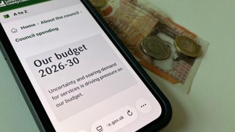A mobile phone showing Leicestershire County Council's budget page and some money