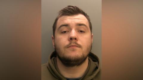 A mugshot of Connor Saggers. He has short brown hair and beard. He has brown eyes and is wearing a olive green hoodie. He is looking at the camera with a serious but somewhat vacant expression.