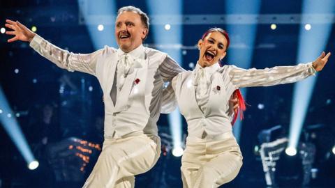 Chris McCausland and Dianne Buswell dancing on Strictly in matching white suits, smiling and arm in arm, each with their other arm outstretched and with a leg raised.