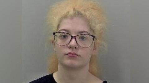 A mugshot of a woman with curly yellow-blonde hair and dark framed glasses. She is wearing a black t-shirt and staring at the camera with a serious expression.