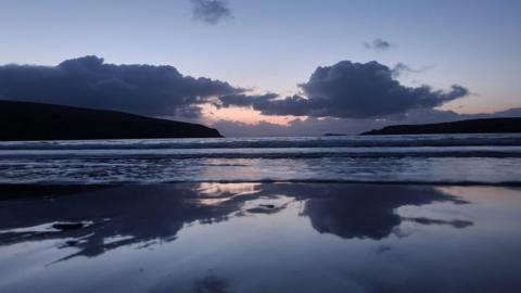 A light blue sky, with some darker clouds and an orange centre point where the sun is rising, sits above mountains and the scene is mirrored in the water below