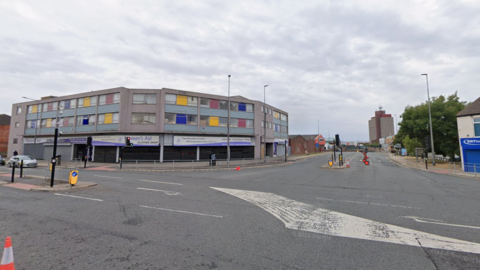 A four-way junction with large white road markings. A large building with colourful markings on the windows can be seen in the distance. The sky is overcast.