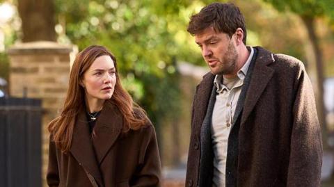 Holliday Grainger acting as Robin Ellacott and Tom Burke as Cormoran Strike in BBC drama Strike. Both actors are walking outside and are wearing brown coats. They are not looking at the camera. 