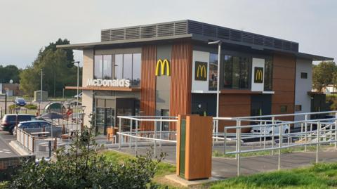 A McDonald's restaurant. It has two storeys. The McDonald's sign is on the front, with three yellow "M" signs to the right. The building is mid-brown wood and white with big glass windows and a dark grey roof. In front of the building is a sloped walkway with silver metal railings.